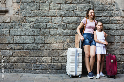 Young happy cheerful positive woman and little girl traveler with suitcase leaning against stone wall