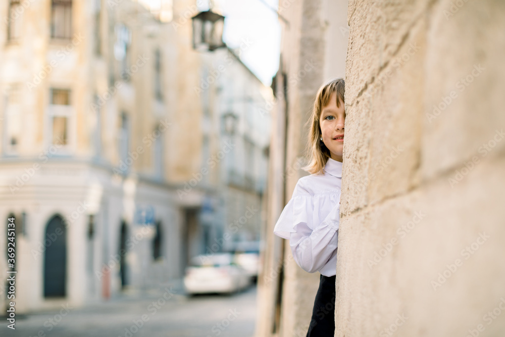 Cute little Caucasian blond girl in black and white clothes, posing to camera outdoors in the old city street, hiding her face behind the ancient building wall Cute little Caucasian blond girl in black and white clothes, posing to camera outdoors in the old city street, hiding her face behind the ancient building wall