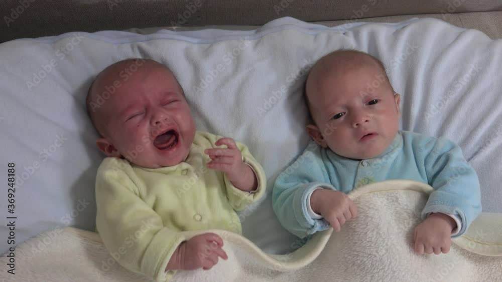 Two newborn girls sisters lie on the bed. Two baby twins on the bed, one crying. Two newborn girls sisters lie on the bed. Two baby twins on the bed, one crying.