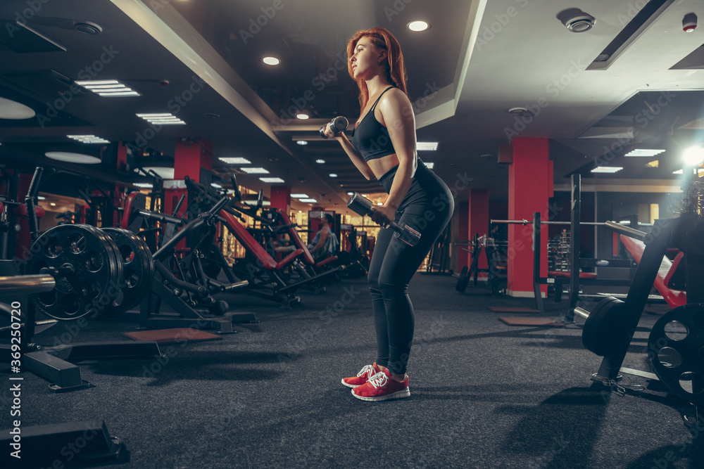 Fototapeta premium Concentrate. Young muscular caucasian woman practicing in gym with equipment. Athletic female model exercising, training her lower body, working out with weights. Wellness, healthy lifestyle
