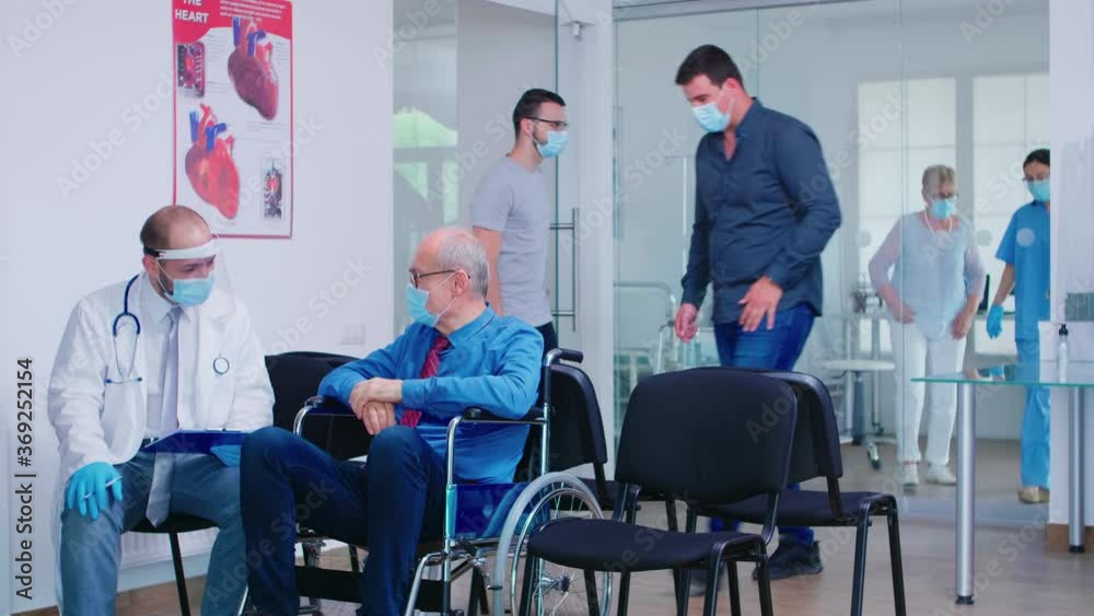 Invalid old man with face mask against infection with coronavirus in wheelchair discussing with doctor in hospital waiting area. Nursing inviting patient in examination room. Invalid old man with face mask against infection with coronavirus in wheelchair discussing with doctor in hospital waiting area. Nursing inviting patient in examination room.