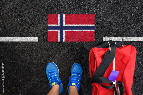 a man with a shoes and travel bag is standing on asphalt next to flag of Norway  and border