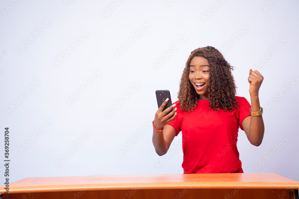 beautiful young black lady sitting at a desk, holding her phone and feeling excited beautiful young black lady sitting at a desk, holding her phone and feeling excited