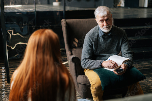 Over the shoulder shot of red-haired young woman patient speaking with mature man psychologist. Skilled psychotherapist asking questions and writing on clipboard during counseling session 