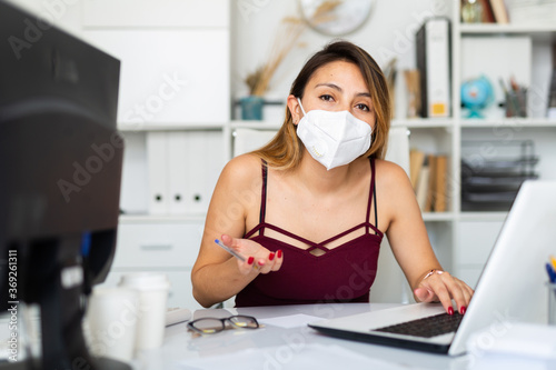 Positive latina woman in protective mask and casual wear working alone with laptop and papers in office