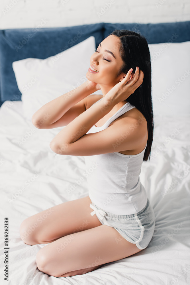 Smiling asian girl touching hair while sitting on bed Smiling asian girl touching hair while sitting on bed