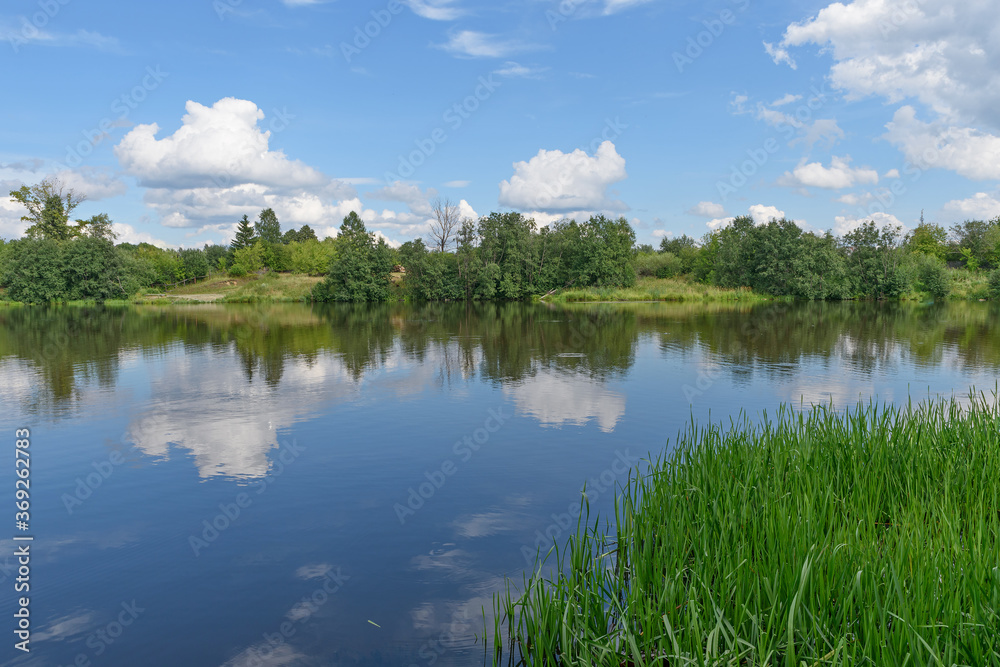 On a summer day the surface of the water of a small river with green banks reflects the blue sky with white clouds