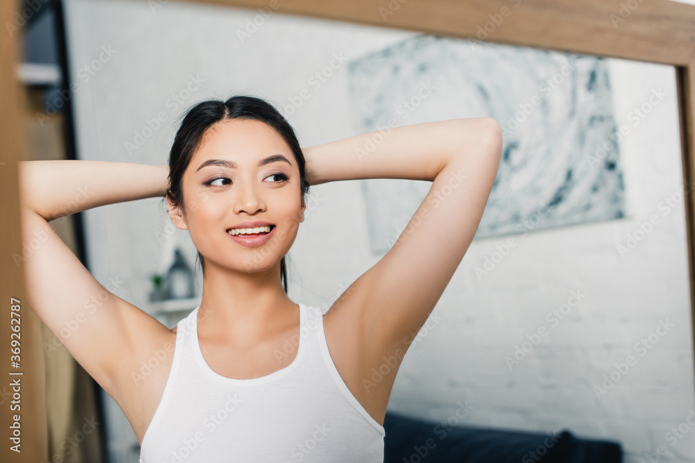 Selective focus of smiling asian woman with hands near head looking at mirror at home Selective focus of smiling asian woman with hands near head looking at mirror at home