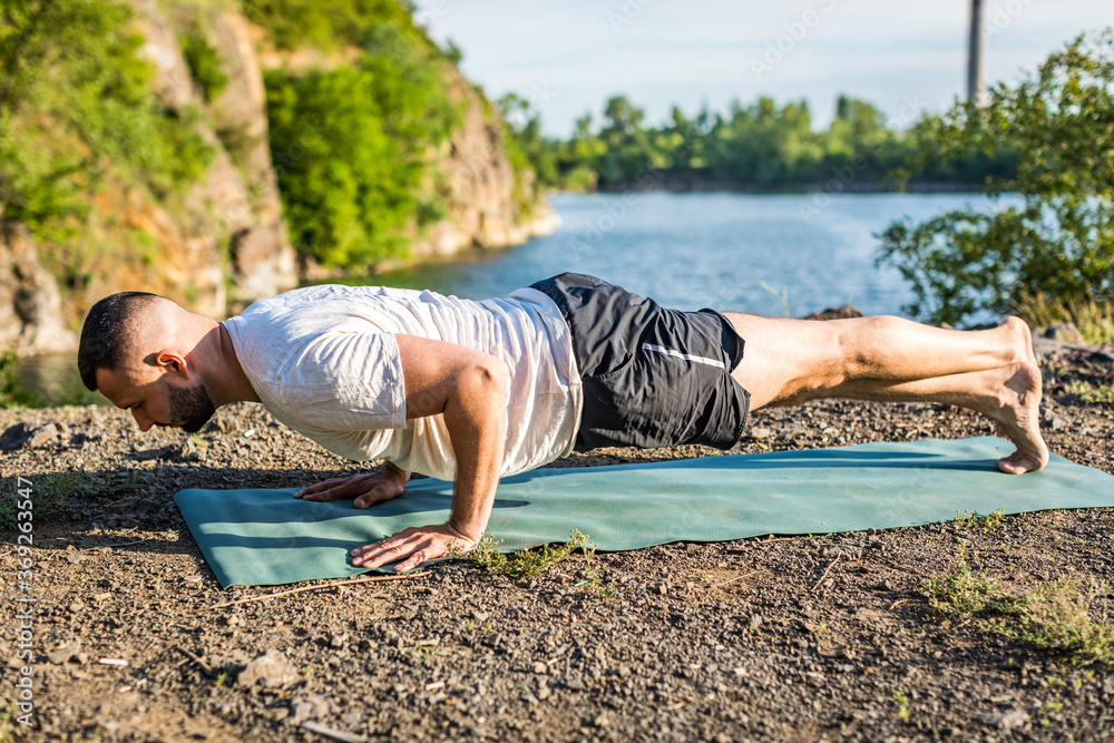 Fototapeta premium Morning and evening, a handsome young man doing yoga exercise on the shore of a quarry lake in nature in the summer outdoors. Healthy active lifestyle concept.