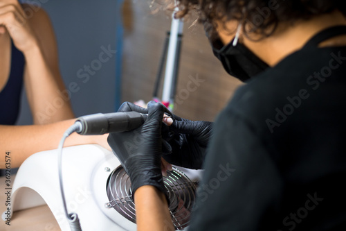 manicurist master in black gloves and mask is making manicure with a manicure drill apparatus in the nail salon