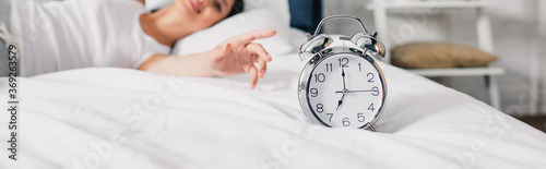 Horizontal crop of alarm clock and smiling asian woman pulling hand on bed at morning