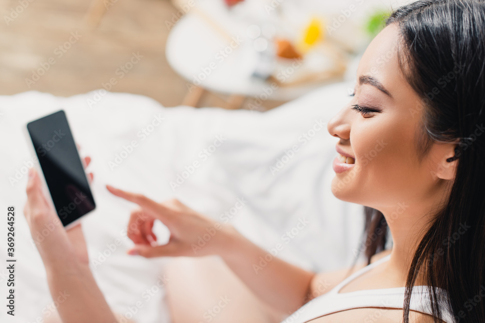 Selective focus of smiling asian girl pointing with finger at smartphone on bed Selective focus of smiling asian girl pointing with finger at smartphone on bed