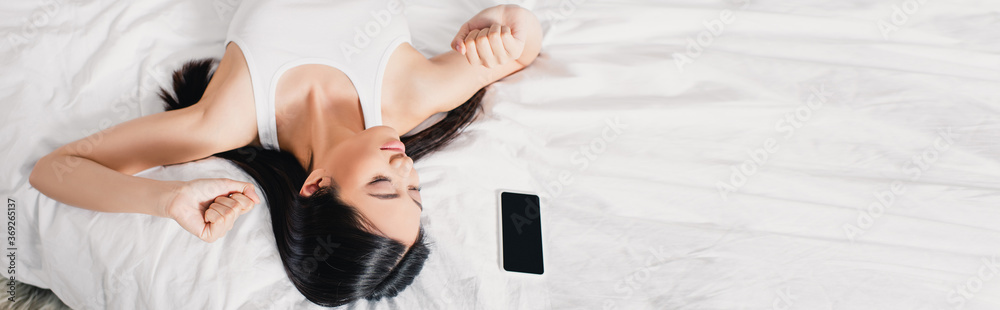 © LIGHTFIELD STUDIOS - Panoramic shot of asian girl stretching near smartphone on bed at morning