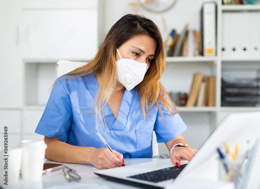 Portrait of young latina female doctor in face mask working on laptop in clinic office