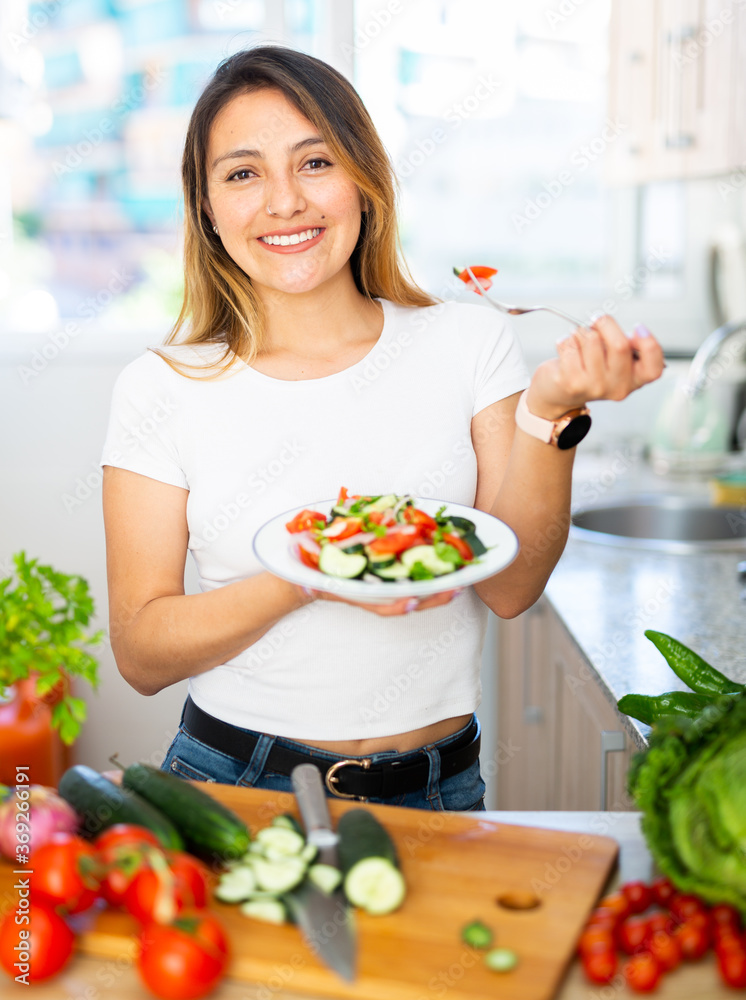 Young mexican housewoman eating vegetable salad from plate at home kitchen