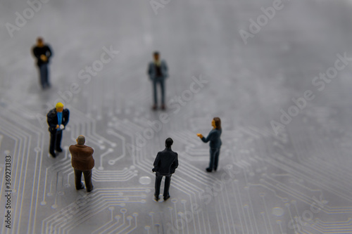 Miniature figurines posed as business people in a meeting over abstract futuristic circuit board, future business concept