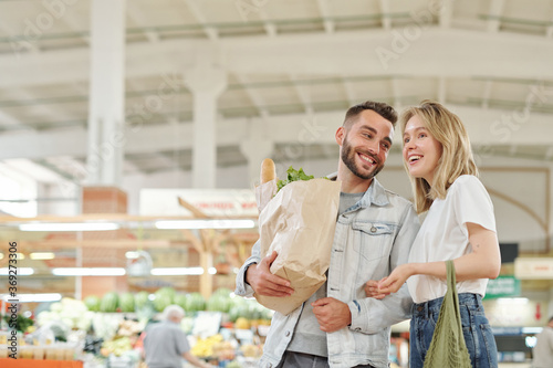 Cheerful young couple standing at farmers market and chatting while enjoying shopping