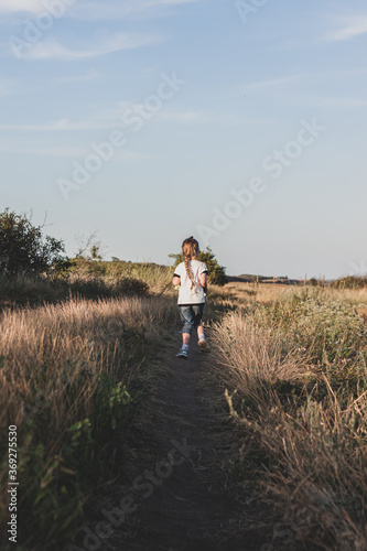 Adorable little girl with long braid running down the hill back view. Happy child running in wild grass countryside landscape. Family walking on fresh air. Local hiking travel concept.