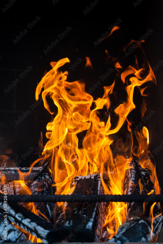 Obraz premium The charred wood in the stove in bright tongues of orange, yellow and red flames against the black background of the smoky wall behind the iron rods of the fireplace.