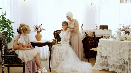 People. Women of different ages sit at an vintage table. They talk and laugh. Senior woman dress family necklaces to the bride. Retro style of the room