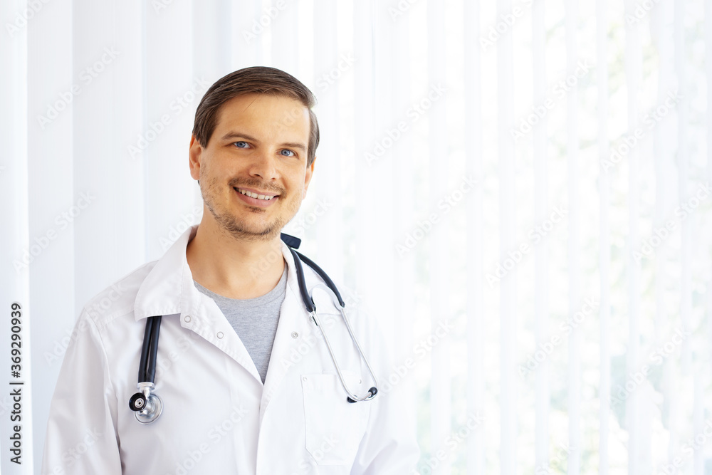 young male doctor of european appearance in white uniform smiling on window background young male doctor of european appearance in white uniform smiling on window background