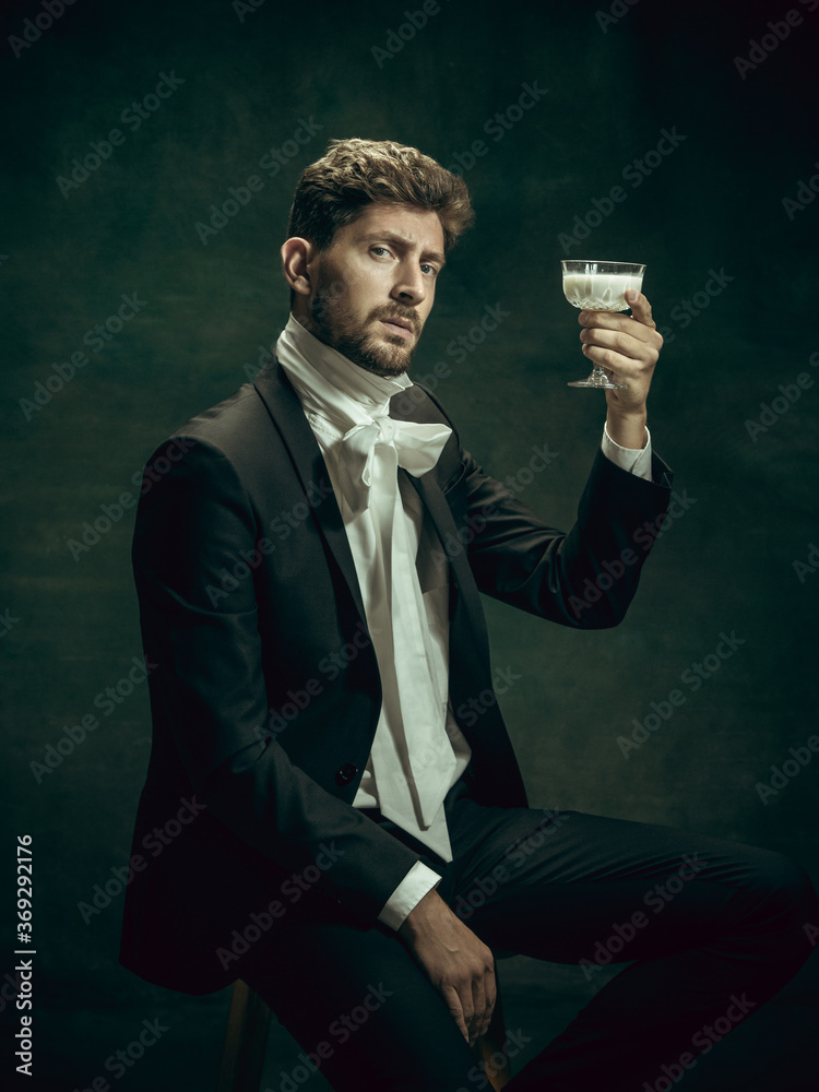 Drinking liquor. Young man in suit as Dorian Gray isolated on dark green background. Retro style, comparison of eras concept. Beautiful male model like classic literature character, old-fashioned. - obrazy, fototapety, plakaty 