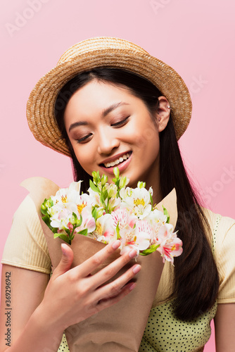young asian woman in straw hat smiling and holding flowers isolated on pink