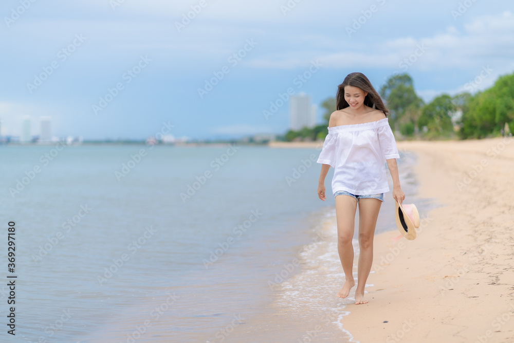 Happy traveler asian woman in white casual wear holding a hat while enjoying a walk on the sea beach at Pattaya, Thailand. Summer travel and vacation concept,