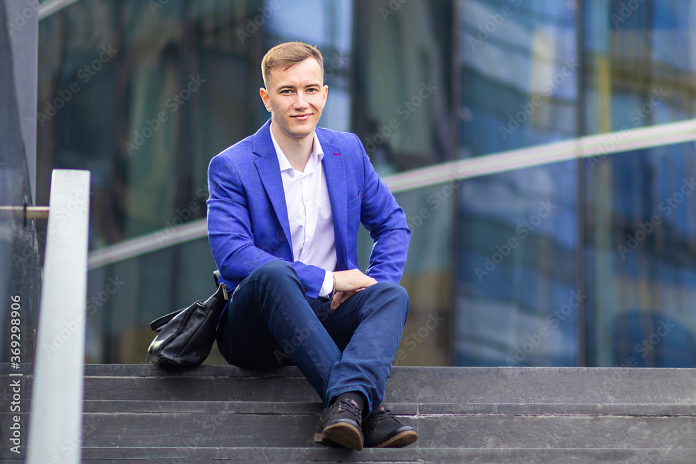 Successful confident businessman sitting on stairs, looking at camera and smiling. Young European manager, office worker in suit outdoors business center. Successful confident businessman sitting on stairs, looking at camera and smiling. Young European manager, office worker in suit outdoors business center.