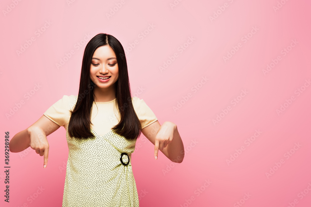 cheerful asian girl pointing with fingers and looking down isolated on pink cheerful asian girl pointing with fingers and looking down isolated on pink