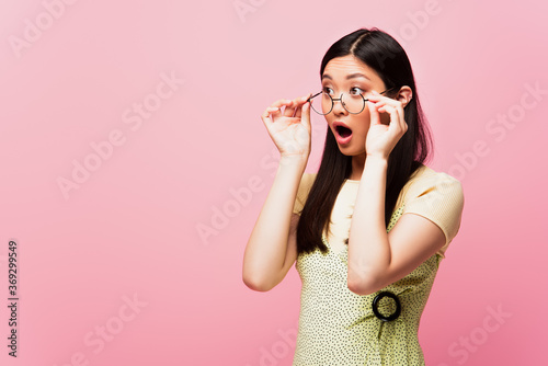 shocked asian girl with open mouth looking away and touching glasses isolated on pink
