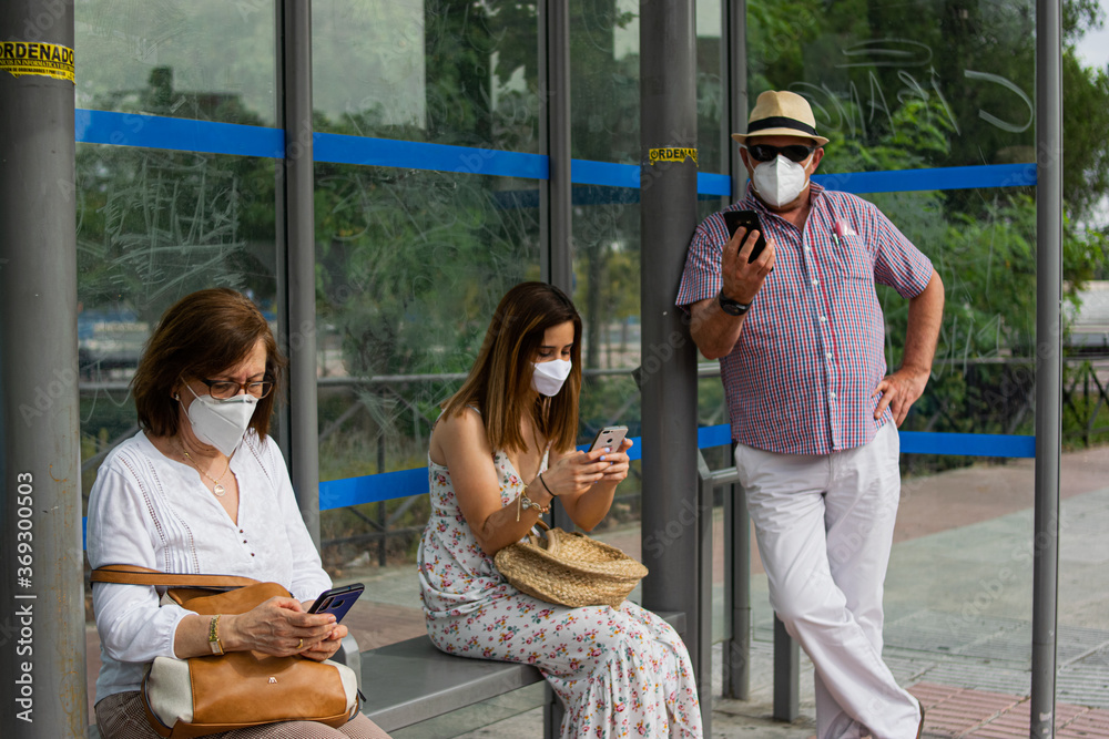 Photo of a three people waiting for the bus checking her phone in a bus stop wearing a white face mask and keeping social distance