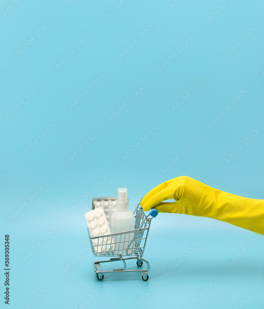 Fototapeta premium Female hand in a yellow medical glove holds a toy shopping cart with medicines on a blue background. Hygiene in the store. Coronavirus pandemic, covid-19.