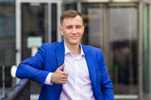 Handsome confident positive man, successful businessman in formal suit, jacket showing thumb up, like, approval gesture. Young European blonde guy looking at camera, smiling outdoors business center