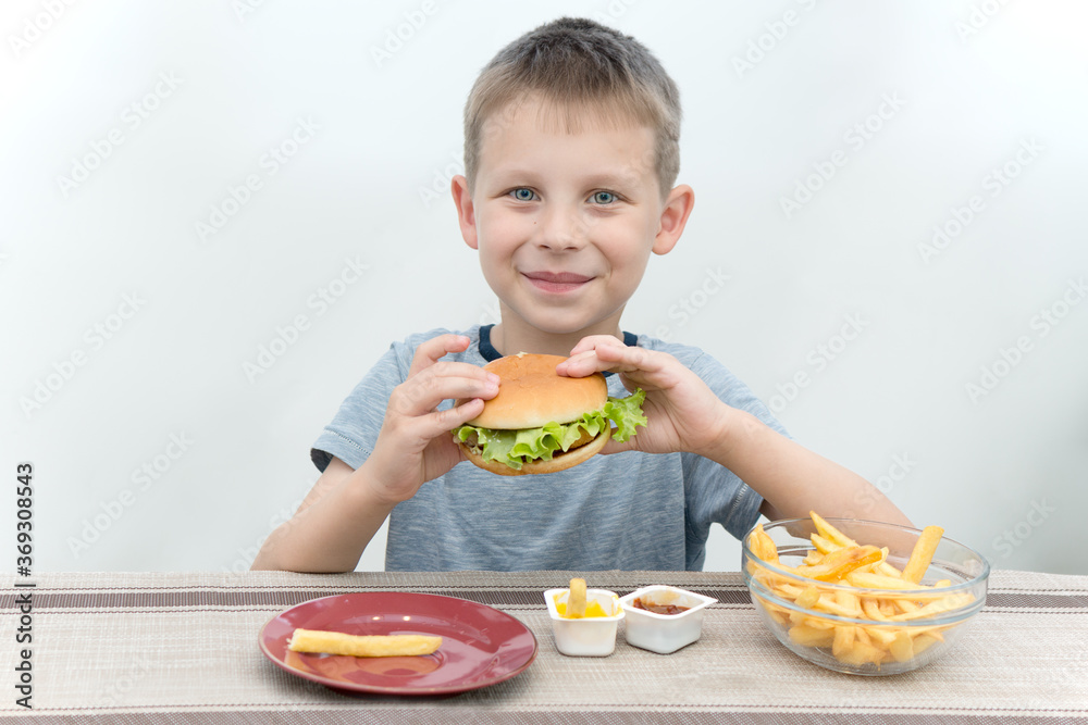 the boy is sitting at the table and holding a fresh hamburger. Looks smiling, fast food on the table.A concept of nutrition that does not bring health benefits, but is very popular with children.