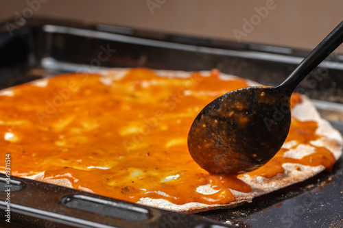 Cooking pizza, spread tomato sauce on a flatbread with a black spoon, close-up