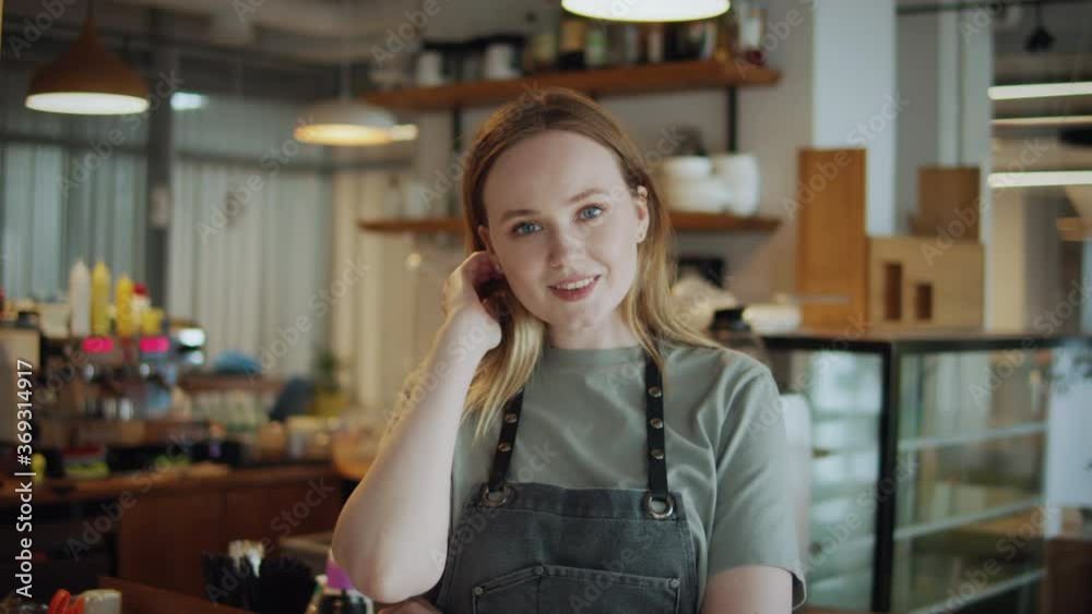 Portrait of happy woman standing in her cafe coffee shop. Cheerful young waitress waiting for clients at coffee shop. Successful small business owner in casual wearing grey apron standing at entrance Portrait of happy woman standing in her cafe coffee shop. Cheerful young waitress waiting for clients at coffee shop. Successful small business owner in casual wearing grey apron standing at entrance