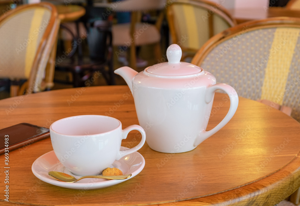 White porcelain teapot, tea cup and smartphone stand on a table in a cafe. Horizontal orientation, selective focus. White porcelain teapot, tea cup and smartphone stand on a table in a cafe. Horizontal orientation, selective focus.
