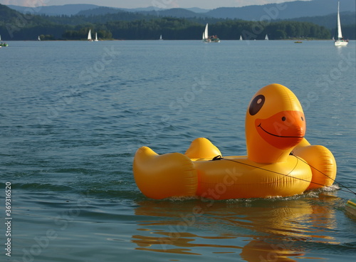 Fototapeta Naklejka Na Ścianę i Meble -  Dmuchana pomarańczowa kaczka do pływania, na  jeziorze, w oddali jachty, łódki, wzgórza otaczające jezioro, wakayjny, letni wypoczynek