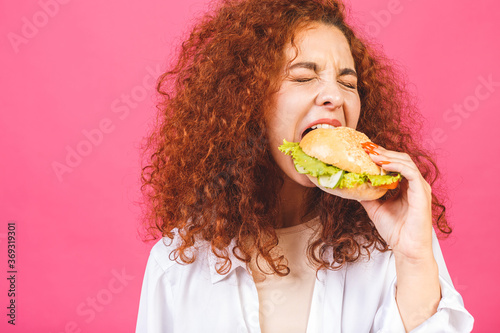 Junk food concept. Attractive Young curly Caucasian Woman Holding And Eating A Large Hamburger  Isolated over pink background.