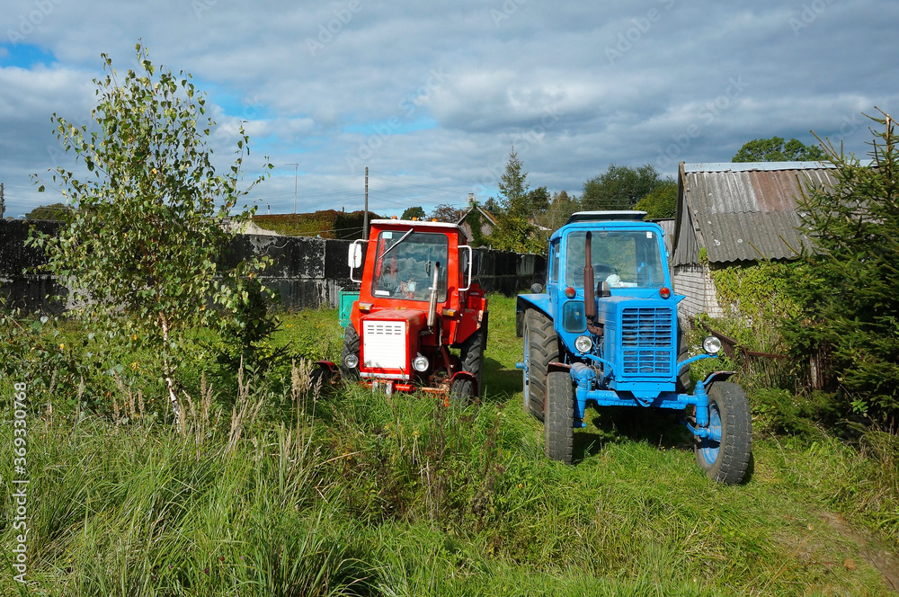 Two tractors in a village near the wood house. Red and blue tractor are in the yard. Two tractors in a village near the wood house. Red and blue tractor are in the yard.