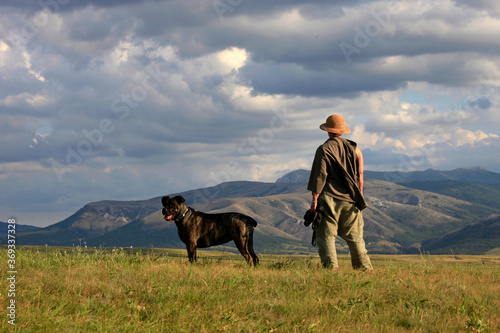 A woman in a hat with a dog of the Cane Corso breed stands on a meadow in a beautiful landscape with a view of the mountains and clouds on a summer day © yanakoroleva27