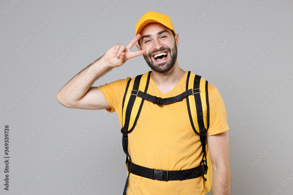 Delivery employee man guy male 20s in yellow cap t-shirt uniform thermal food bag backpack work courier service during quarantine coronavirus covid-19 virus, posing isolated on gray background studio. Delivery employee man guy male 20s in yellow cap t-shirt uniform thermal food bag backpack work courier service during quarantine coronavirus covid-19 virus, posing isolated on gray background studio.