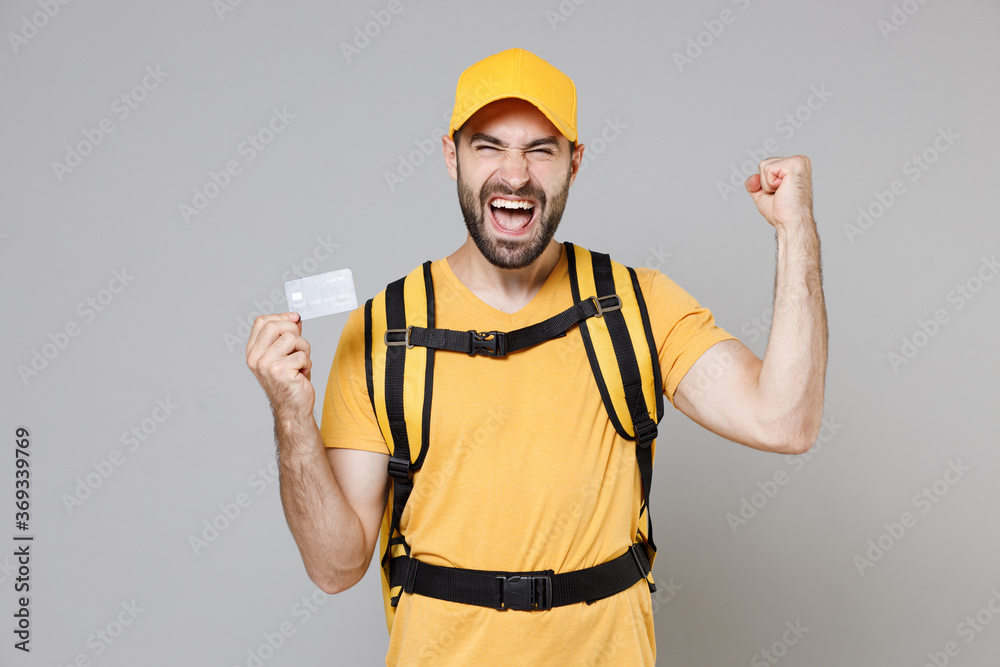 Delivery employee man guy male 20s in yellow cap t-shirt uniform thermal food bag backpack work courier service on quarantine covid-19 hold in hand bank credit card isolated on gray background studio.
