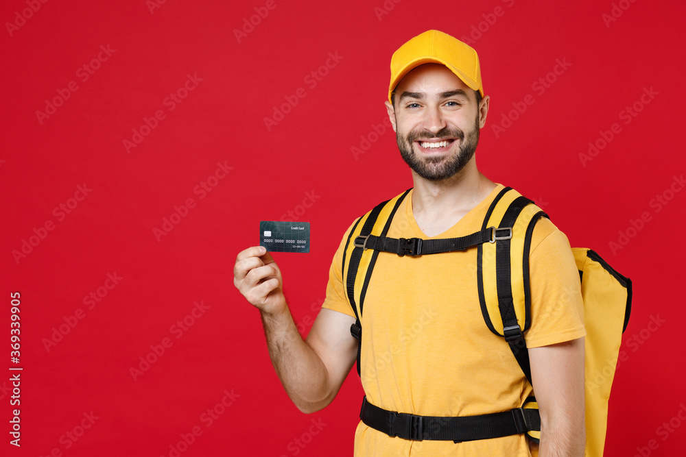 Delivery man in yellow cap t-shirt uniform thermal bag backpack with food hold credit card isolated on red background studio Guy male employee work courier Service coronavirus covid-19 virus concept Delivery man in yellow cap t-shirt uniform thermal bag backpack with food hold credit card isolated on red background studio Guy male employee work courier Service coronavirus covid-19 virus concept