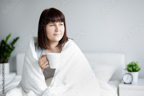 young woman wrapped in blanket sitting in bed, drinking coffee and thinking or dreaming about something at home