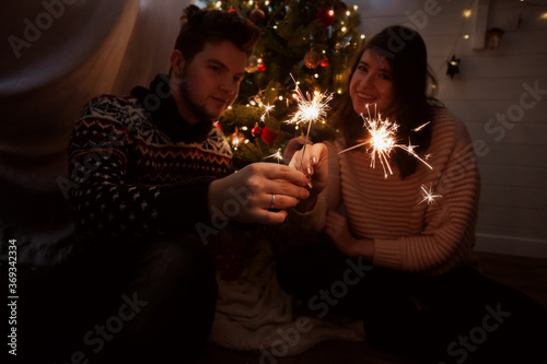 Happy couple holding fireworks under christmas tree with lights. Young family with burning sparklers celebrating together in festive dark room. Happy New Year