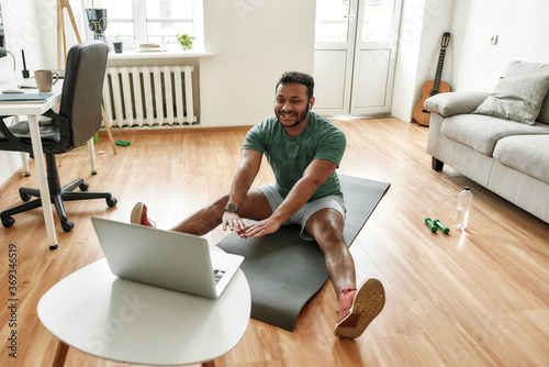 Cheerful stretching. Male fitness instructor showing exercises while streaming, broadcasting video lesson on training at home using laptop. Sport, online gym concept