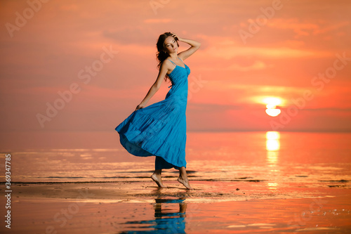 Young romantic girl walks relaxed by the water at sunset or sunrise. Beautiful woman in blue dress in boho style on background at sea. Sunrise over the ocean. Summertime scene