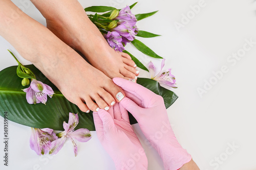 Fototapeta Naklejka Na Ścianę i Meble -  Beautiful perfect skin female legs feet top view with tropical flowers and green palm leaf. Hands of beautician in pink gloves. Nail polish, care and clean, spa pedicure treatment in white. Copy space